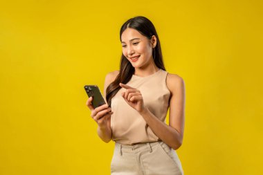 İzole edilmiş sarı arka planda cep telefonu kullanan Asyalı güzel kadın. Çekici genç kız manken stüdyo ekranında dijital akıllı telefon cihazlarıyla sohbet ederken mutlu ve neşeli hisseder..