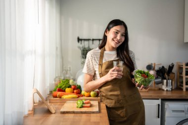 Asyalı kadın portresi sağlıklı salata yiyor ve temiz su içiyor. Önlük giyen güzel bir kız, sabahları evde sağlık hizmetleri için güzel yemekler yapmaktan zevk alıyor ve gülümsüyor, kameraya bakıyor..