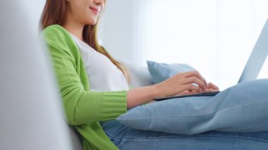 Asyalı genç bir kadın dizüstü bilgisayar kullanıyor ve evden çalışıyor. Çekici, serbest çalışan güzel bir kadın. Kanepede oturuyor. Oturma odasında dizüstü bilgisayar kullanıyor.