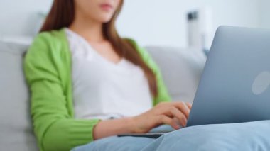 Asyalı genç bir kadın dizüstü bilgisayar kullanıyor ve evden çalışıyor. Çekici, serbest çalışan güzel bir kadın. Kanepede oturuyor. Oturma odasında dizüstü bilgisayar kullanıyor.