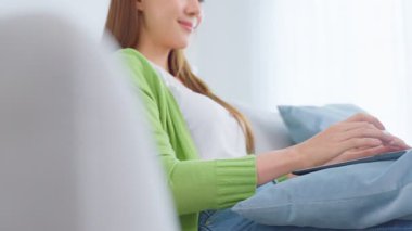 Asyalı genç bir kadın dizüstü bilgisayar kullanıyor ve evden çalışıyor. Çekici, serbest çalışan güzel bir kadın. Kanepede oturuyor. Oturma odasında dizüstü bilgisayar kullanıyor.