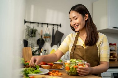 Asyalı genç bir kadın sabah evde mutfakta sağlıklı yemekler pişiriyor. Çekici kadın ev hanımı kendini mutlu ve rahat hissediyor, malzemeleri hazırlamaktan ve kahvaltıda yeşil salata yapmaktan hoşlanıyor..
