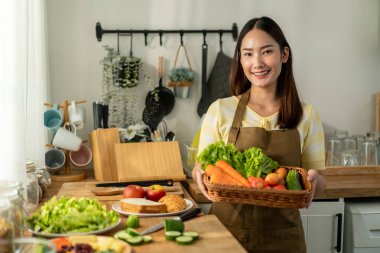 Sabahları evde, mutfakta sağlıklı yemek pişiren kadın portresi. Çekici kadın ev hanımı mutlu ve rahatlamış hisseder. Malzemeleri hazırlarken kameraya bakar ve kahvaltı için yeşil salata yapar.