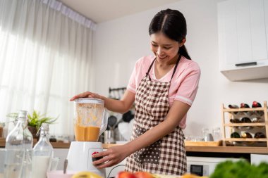Asyalı kadın evdeki mutfakta blender 'da taze meyve suyu yapıyor. Çekici kadın ev hanımı mutlu ve rahat hisseder, malzemeleri hazırlamaktan ve sabah kahvaltısı için sağlıklı içecekler hazırlamaktan zevk alır..