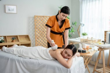 Asyalı genç bir kadın bitkisel poşetlerle Tayland masajı yaptırıyor. Çekici, güzel bir kız masaj yatağında uzanıp masaj salonundaki masözden rahatlatıcı fizyoterapi hizmeti alıyor..