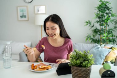 Asyalı genç bir kadın evdeki oturma odasında Amerikan kahvaltısı yiyor. Çekici bir kız masaya oturur, mutlu olur ve rahatlar. Evde tablet cihazla film izlerken İngilizce kahvaltısını sağlık için yer..