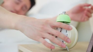 Genç bir yetişkinin eli uzanıyor, çalar saati kapatıyor, yatakta uyanıyor..