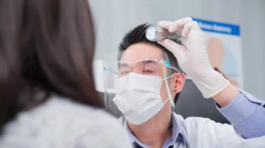 Maskeli, gözlüklü, eldivenli Asyalı adam tıbbi düzende şişeyi dikkatle tutuyor..