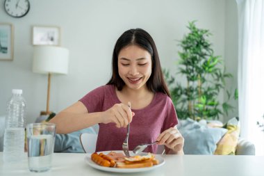 Asyalı genç bir kadın evdeki oturma odasında Amerikan kahvaltısı yiyor. Çekici kız masaya oturur, sabahları evde sağlık için temiz suyla İngiliz kahvaltısı yapar. Diyet ve Sağlıklı Yemek Konsepti.