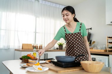 Asyalı genç bir kadın sabahları mutfakta sağlıklı yemek pişiriyor. Önlük giyen çekici bir kız kendini mutlu ve rahat hisset, evde tava kullanarak kahvaltı hazırlamak için gerekli malzemeleri hazırlarken keyfine bak..