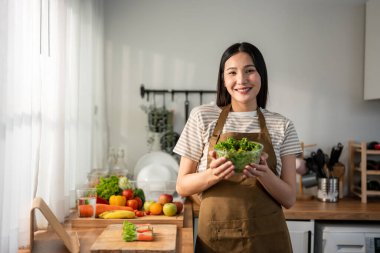 Asyalı kadın portresi evde mutfakta yeşil sağlıklı salata pişiriyor. Önlük giyen güzel bir genç kız, sabahları evdeki sağlık hizmetleri için temiz yemek yapmaktan zevk alıyor ve gülümsüyor, kameraya bakıyor..