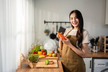 Asyalı kadın portresi evde mutfakta yeşil sağlıklı salata pişiriyor. Önlük giyen güzel bir genç kız, sabahları evdeki sağlık hizmetleri için temiz yemek yapmaktan zevk alıyor ve gülümsüyor, kameraya bakıyor..