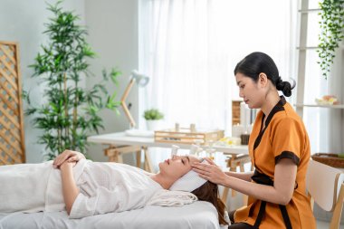 Asyalı genç bir kadın güzellik merkezinde kafa ve yüz masajı yaptırıyor. Çekici kız masaj yatağında uzanıyor, masaj kliniğinde vücut bakımı için masözden rahatlatıcı fizyoterapi hizmeti alıyor..