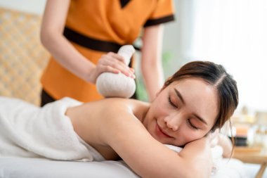 Asyalı genç bir kadın bitkisel poşetlerle Tayland masajı yaptırıyor. Çekici, güzel bir kız masaj yatağında uzanıp masaj salonundaki masözden rahatlatıcı fizyoterapi hizmeti alıyor..