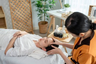 Asyalı genç bir kadın güzellik merkezinde kafa ve yüz masajı yaptırıyor. Çekici kız masaj yatağında uzanıyor, masaj kliniğinde vücut bakımı için masözden rahatlatıcı fizyoterapi hizmeti alıyor..