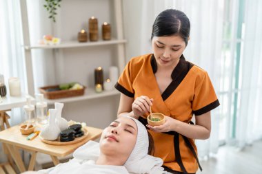 Asyalı genç bir kadın maske takarken mutlu ve rahat hisseder. Çekici güzel kız masaj masasında uzanıyor, güzellik salonundaki cilt ve vücut bakımı için masözden fizyoterapi alıyor..