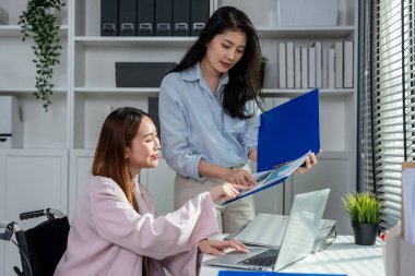 Tekerlekli sandalyedeki Asyalı iş kadını ofiste iş arkadaşıyla çalışıyor. Çekici genç bayan çalışan mutlu ve rahatlamış hissediyor, şirket işyerinde dizüstü bilgisayarla proje tartışmaktan zevk alıyor.