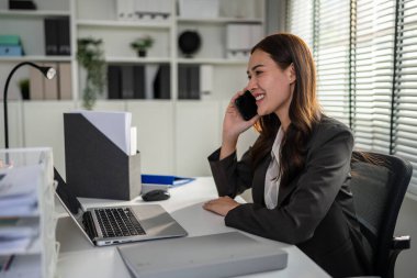 Asyalı genç iş kadını akıllı telefondan konuşuyor ve ofiste çalışıyor. Çekici bayan çalışan masada oturuyor, iş yerini planlamak için meslektaşıyla cep telefonu görüşmesi yapıyor..