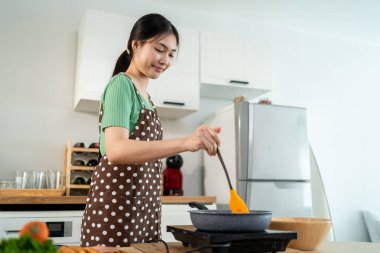 Asyalı genç bir kadın sabahları mutfakta sağlıklı yemek pişiriyor. Önlük giyen çekici bir kız kendini mutlu ve rahat hisset, evde tava kullanarak kahvaltı hazırlamak için gerekli malzemeleri hazırlarken keyfine bak..