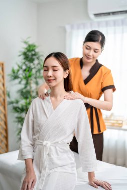 Asyalı güzel bir kadın güzellik merkezinde omuz ve sırt masajı yaptırıyor. Masaj yatağında oturan çekici genç kız, masaj kliniğinde vücut bakımı için masözden rahatlatıcı fizyoterapi hizmeti alıyor..