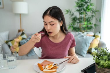 Asyalı genç bir kadın evdeki oturma odasında Amerikan kahvaltısı yiyor. Çekici kız masaya oturur, sabahları evde sağlık için temiz suyla İngiliz kahvaltısı yapar. Diyet ve Sağlıklı Yemek Konsepti.