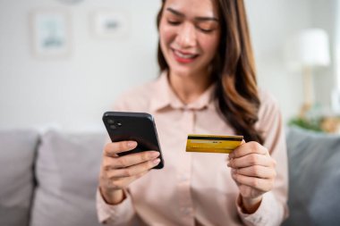 Evdeki oturma odasında cep telefonu alışverişi yapan Asyalı bir kadın. Çekici güzel bir kız, elinde kredi kartıyla kanepeye oturur evdeki web mağazasında akıllı telefonlardan alışveriş yaptıktan sonra ödeme yapar.