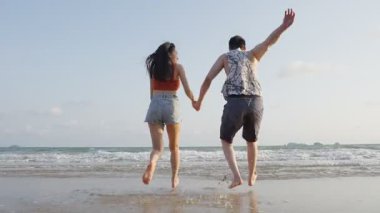 Sahilde el ele tutuşan genç çiftin arka görüntüsü.