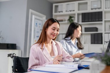 Tekerlekli sandalyede oturan Asyalı iş kadını ve ofiste masa başında çalışan. Çekici genç bayan çalışan ekibi mutlu ve şirket işyerindeki bilgisayar tartışması projesini kullanarak eğleniyor..