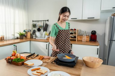 Asyalı genç bir kadın sabahları mutfakta sağlıklı yemek pişiriyor. Önlük giyen çekici bir kız kendini mutlu ve rahat hisset, evde tava kullanarak kahvaltı hazırlamak için gerekli malzemeleri hazırlarken keyfine bak..