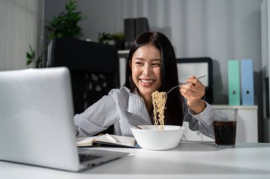 Ofiste çalışırken erişte yiyen Asyalı genç iş kadını. Çekici profesyonel bayan çalışan masada oturuyor, aç ve aç hissediyor sonra da şirket işyerinde yemek yiyor..