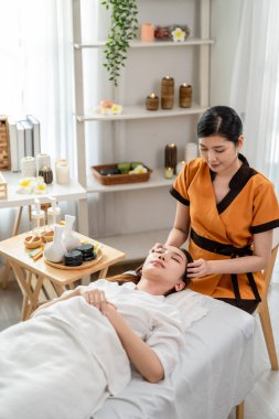 Asyalı genç bir kadın güzellik merkezinde kafa ve yüz masajı yaptırıyor. Çekici kız masaj yatağında uzanıyor, masaj kliniğinde vücut bakımı için masözden rahatlatıcı fizyoterapi hizmeti alıyor..