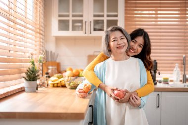 Evde kızıyla birlikte duran Asyalı yaşlı anne portresi. Çekici torun ve büyükanne mutlu ve rahatlamış hissediyorlar, sağlıklı meyveler tutuyorlar ve evdeki kameraya bakıyorlar..