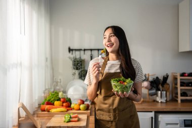 Asyalı çekici kadın evde yeşil sağlıklı salata yiyor. Önlük giyen genç ve güzel bir kız, sabahları evdeki sağlık hizmetleri için temiz yemek yapıyor. Diyet ve Sağlıklı Yemek Konsepti.