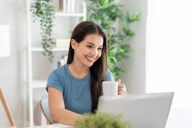 Evdeki oturma odasında dizüstü bilgisayar kullanırken kahve içen Asyalı kadın. Çekici, sıradan bir kız mutlu olur ve boş zamanlarını evde çalışarak geçirir. Etkinlik yaşam tarzları konsepti.