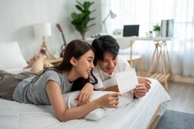 Asyalı genç çift evdeki yatak odasında sevgililer günü posteri okuyor. Çekici, romantik, yeni evli bir erkek ve kadın yatağa uzanıp hafta sonları boş zamanlarını evde geçiriyorlar..