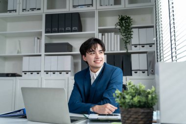 Ofiste çalışan Asyalı çekici iş adamı. Genç profesyonel erkek işçi mutlu ve rahat hissediyor. Şirket bölümündeki dizüstü bilgisayar planı projesini kullanarak masada oturuyor..