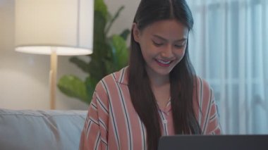 Genç Asyalı kadın kanepede oturup gülümserken dizüstü bilgisayar kullanıyor.