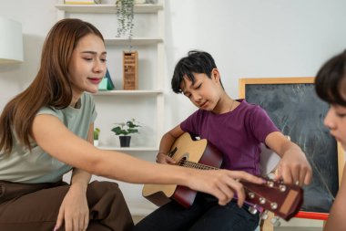 Asyalı öğretmen ilkokulda gitar sınıfında öğrencileri eğitiyor. Hevesli kadın öğretmen, sandalyede oturan ve dikkatle gitar çalan sevimli çocuklara dersi açıklıyor..