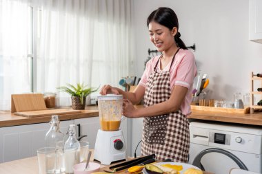 Asyalı kadın evdeki mutfakta blender 'da taze meyve suyu yapıyor. Çekici kadın ev hanımı mutlu ve rahat hisseder, malzemeleri hazırlamaktan ve sabah kahvaltısı için sağlıklı içecekler hazırlamaktan zevk alır..
