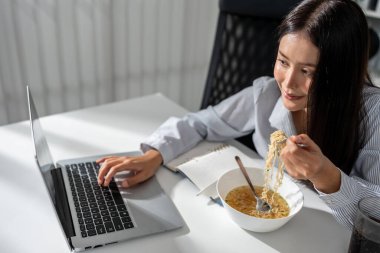 Ofiste çalışırken erişte yiyen Asyalı genç iş kadını. Çekici profesyonel bayan çalışan masada oturuyor, aç ve aç hissediyor sonra da şirket işyerinde yemek yiyor..