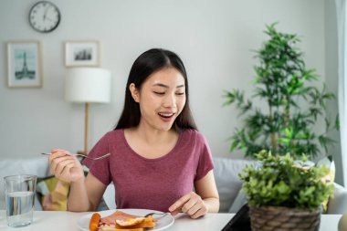 Asyalı genç bir kadın evdeki oturma odasında Amerikan kahvaltısı yiyor. Çekici bir kız masaya oturur, mutlu olur ve rahatlar. Evde tablet cihazla film izlerken İngilizce kahvaltısını sağlık için yer..