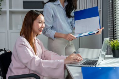 Tekerlekli sandalyedeki Asyalı iş kadını ofiste iş arkadaşıyla çalışıyor. Çekici genç bayan çalışan mutlu ve rahatlamış hissediyor, şirket işyerinde dizüstü bilgisayarla proje tartışmaktan zevk alıyor.