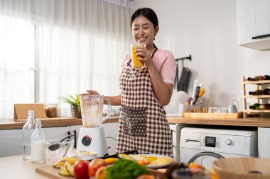 Asyalı kadın evdeki mutfakta blender 'da taze meyve suyu yapıyor. Çekici kadın ev hanımı mutlu ve rahat hisseder, malzemeleri hazırlamaktan ve sabah kahvaltısı için sağlıklı içecekler hazırlamaktan zevk alır..