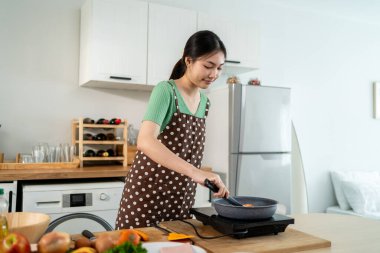 Asyalı genç bir kadın sabahları mutfakta sağlıklı yemek pişiriyor. Önlük giyen çekici bir kız kendini mutlu ve rahat hisset, evde tava kullanarak kahvaltı hazırlamak için gerekli malzemeleri hazırlarken keyfine bak..
