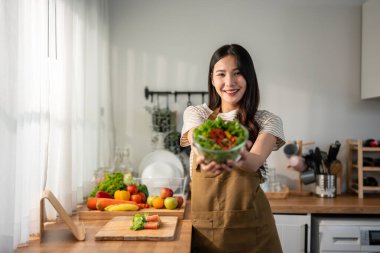 Asyalı kadın portresi evde mutfakta yeşil sağlıklı salata pişiriyor. Önlük giyen güzel bir genç kız, sabahları evdeki sağlık hizmetleri için temiz yemek yapmaktan zevk alıyor ve gülümsüyor, kameraya bakıyor..