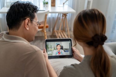 Asyalı olgun bir hasta, tabletteki tıp doktoruyla sanal konuşma yapıyor. Çekici hekim kadın teletıp görüntülü konuşma yaparak evdeki hasta çifte danışmanlık hizmeti veriyor..