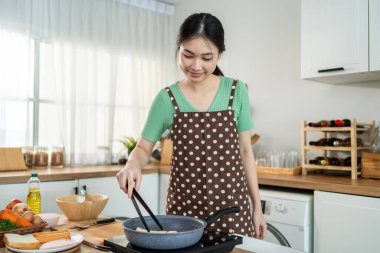 Asyalı genç bir kadın sabahları mutfakta sağlıklı yemek pişiriyor. Önlük giyen çekici bir kız kendini mutlu ve rahat hisset, evde tava kullanarak kahvaltı hazırlamak için gerekli malzemeleri hazırlarken keyfine bak..