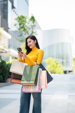 Mağazada beklerken cep telefonu kullanan Asyalı genç bir kadın. Çekici, güzel bir bayan alışveriş çantası tutuyor. Mutlulukla dışarı çıkıyor. Alışveriş merkezinden satın almanın keyfini çıkarıyor..