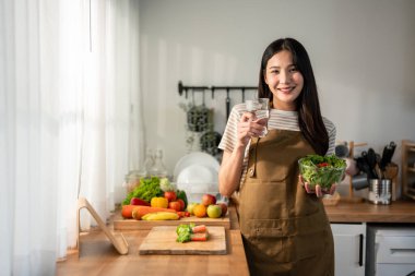 Asyalı kadın portresi sağlıklı salata yiyor ve temiz su içiyor. Önlük giyen güzel bir kız, sabahları evde sağlık hizmetleri için güzel yemekler yapmaktan zevk alıyor ve gülümsüyor, kameraya bakıyor..