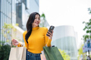 Mağazada beklerken cep telefonu kullanan Asyalı genç bir kadın. Çekici, güzel bir bayan alışveriş torbaları tutuyor. Mutlulukla dışarı çıkıyor. Alışveriş merkezinden satın almanın keyfini çıkarıyor..